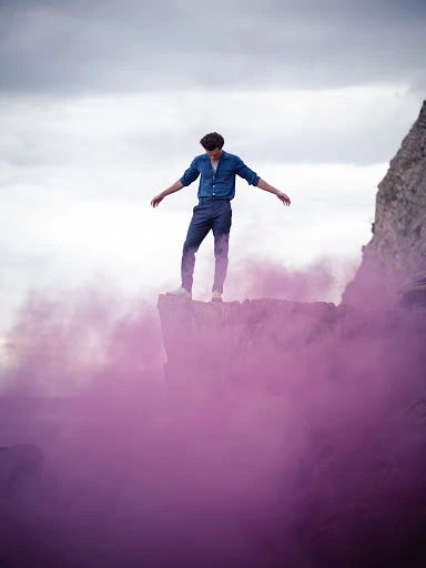 A lone man strikes a pose atop a rocky cliff enveloped in swirling magenta smoke, set against a backdrop of a cloudy sky and coastline.