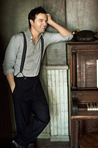 Portrait of a man in a grey shirt and suspenders leaning on an old piano in a warmly lit indoor environment with vintage decor.