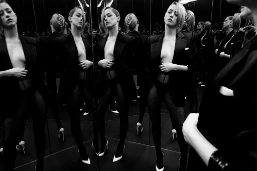 Black and white fashion photograph of a woman posing in a blazer and stockings, reflected in a mirror, creating a repetitive visual effect.