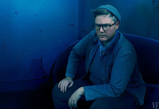 Headshot of a seated adult with short hair, wearing glasses and a beanie, gazing to the side against a distressed blue wall.