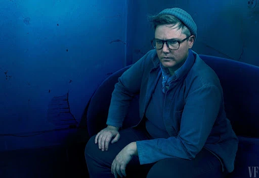 Headshot of a seated adult with short hair, wearing glasses and a beanie, gazing to the side against a distressed blue wall.