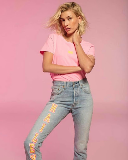 Blonde woman poses in pink t-shirt and jeans with writing down leg, against a pink backdrop, fashion studio shot.