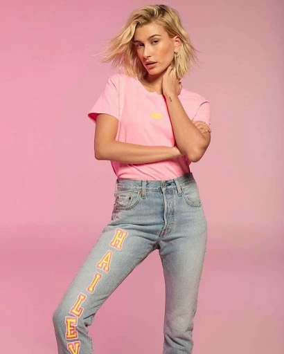 Blonde woman poses in pink t-shirt and jeans with writing down leg, against a pink backdrop, fashion studio shot.