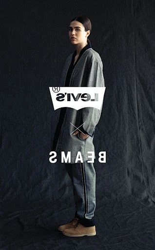 Woman wearing a gray jacket and pants with tan shoes, posing in front of a dark gray background, with Levi's and BEAMS logos displayed.