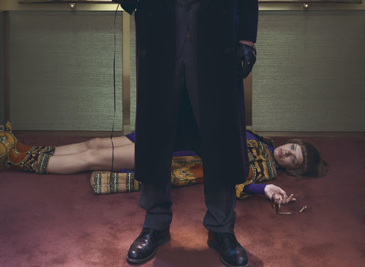 Indoor shot of a woman in a patterned dress lying on the floor while a person in a dark coat stands over her, holding glasses.