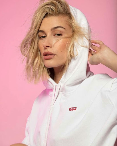 Close-up of a woman in a white hoodie with blonde hair against a pink backdrop in a studio environment, fashion and beauty shot.