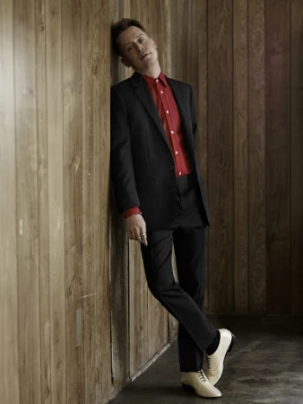 Full-length portrait of a man leaning against a vertical wood-paneled wall, wearing a black suit and red shirt.