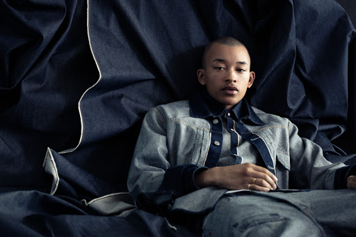 Portrait of a young adult with a shaved head wearing a denim jacket, seated against a dark blue denim fabric backdrop in a studio.