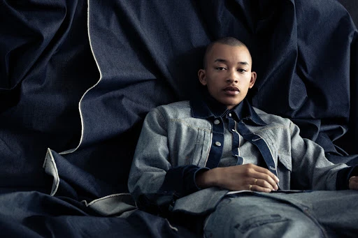 Portrait of a young adult with a shaved head wearing a denim jacket, seated against a dark blue denim fabric backdrop in a studio.