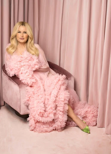 Jennifer Coolidge sits in a pink chair wearing a pink ruffled robe and green heels against a matching pink draped backdrop.