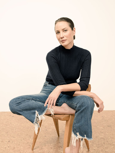Woman with slicked-back hair wearing a ribbed black turtleneck and distressed denim jeans seated on a wooden chair.