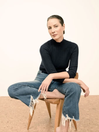 Woman with slicked-back hair wearing a ribbed black turtleneck and distressed denim jeans seated on a wooden chair.