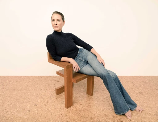 Woman with hair pulled back seated barefoot on a wooden chair wearing a dark ribbed turtleneck and blue denim jeans.