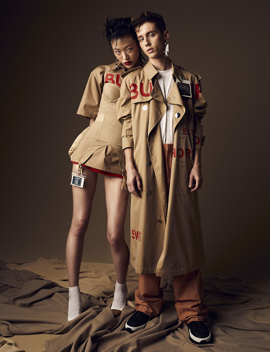 Two fashion models, an Asian woman and a Caucasian man, pose in Burberry outfits with painted logos in a photo studio setting.