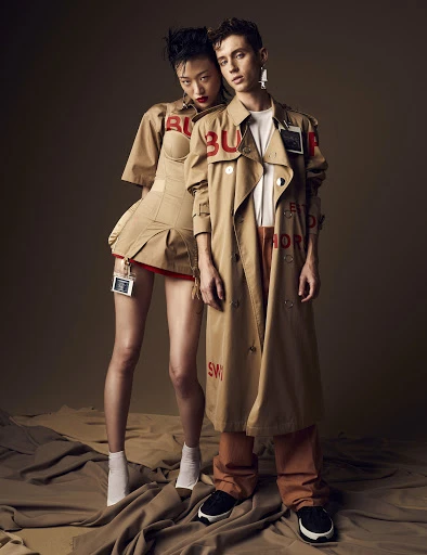 Two fashion models, an Asian woman and a Caucasian man, pose in Burberry outfits with painted logos in a photo studio setting.