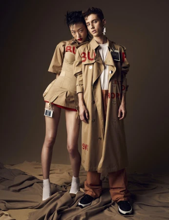 Two fashion models, an Asian woman and a Caucasian man, pose in Burberry outfits with painted logos in a photo studio setting.