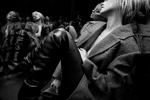 Monochrome fashion shot of a blonde woman in a herringbone coat, shiny tights, and black leather boots posing in a reflective setting.
