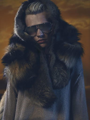 Person wearing sunglasses and a fur-trimmed hood in a studio setting against an orange backdrop with a subtle, enigmatic expression.