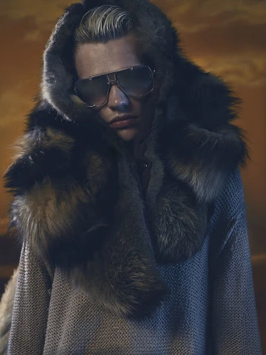 Person wearing sunglasses and a fur-trimmed hood in a studio setting against an orange backdrop with a subtle, enigmatic expression.