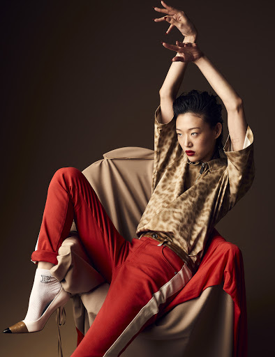 Asian woman in animal print top and red pants sits in a beige chair, raising her arms in a studio setting with a brown background.