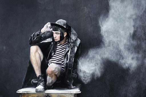 Man poses wearing a black leather jacket and hat, powdered with dust sitting on an ornately carved end table, against a dark backdrop.