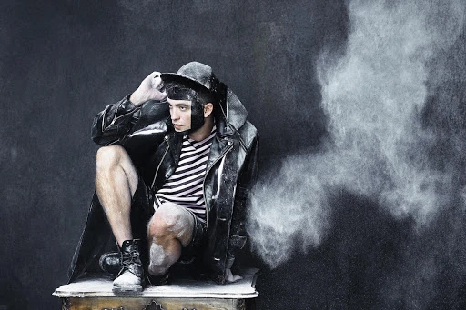 Man poses wearing a black leather jacket and hat, powdered with dust sitting on an ornately carved end table, against a dark backdrop.