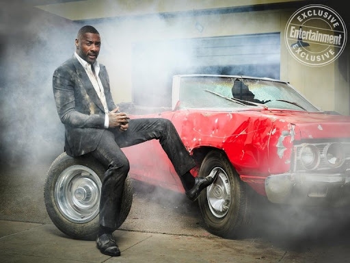 Idris Elba sitting on a tire in a dirty grey suit beside a dented red convertible amid smoke, promoting an Entertainment Weekly exclusive.
