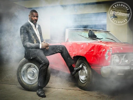 Idris Elba sitting on a tire in a dirty grey suit beside a dented red convertible amid smoke, promoting an Entertainment Weekly exclusive.
