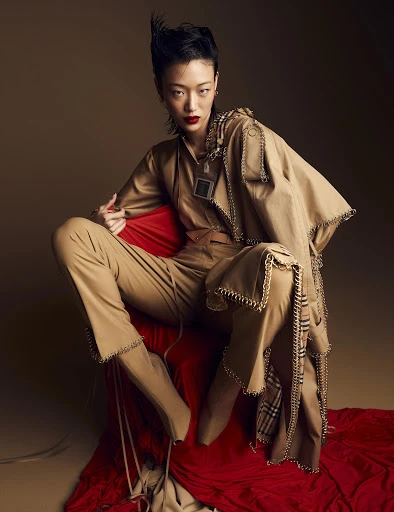 An Asian female model sits on a red draped surface, wearing a tan jumpsuit and trench coat with edgy chain detailing, against a studio backdrop.
