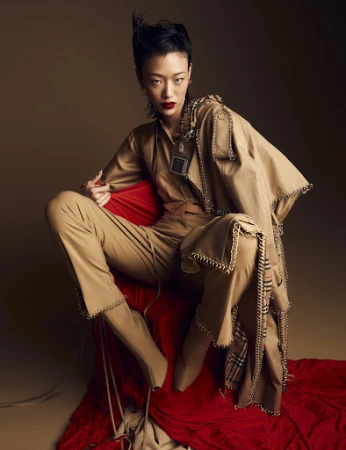 An Asian female model sits on a red draped surface, wearing a tan jumpsuit and trench coat with edgy chain detailing, against a studio backdrop.