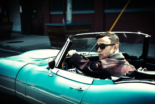 Stylish adult male driving a teal convertible car on a city street, wearing sunglasses and a patterned jacket, captured in outdoor setting.