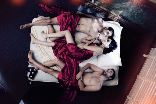 Overhead shot of two men and one woman lying on a mattress draped in red satin fabric indoors, with light streaming through a window.