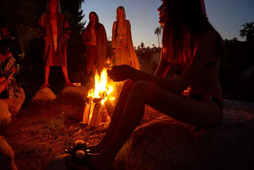A group of people are gathered around a bonfire at dusk, silhouetted against the twilight sky with palm trees in the distance.