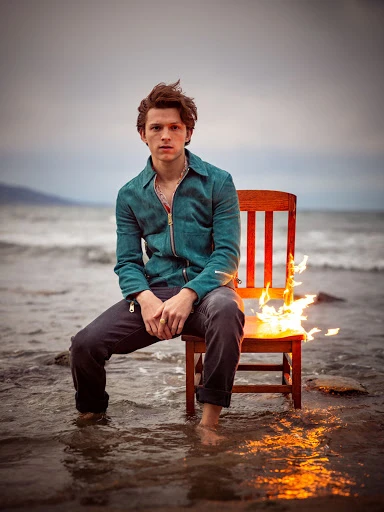 A man sits on a burning chair on a beach, wearing a zip-up shirt, dark pants, and no shoes, with the ocean and sky visible in the background.