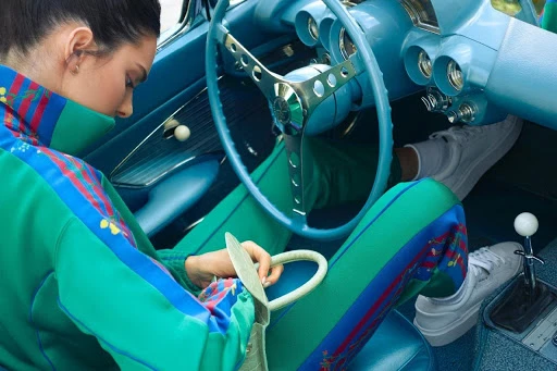Woman wearing a green tracksuit seated inside a vintage blue car holding a light green handbag