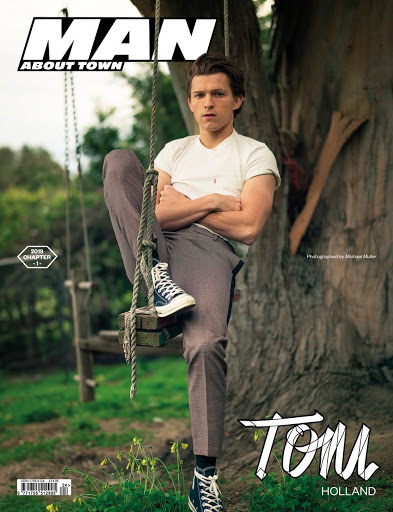 Tom Holland poses outdoors wearing a white t-shirt and grey pants, seated on a wooden swing hanging from a mature tree, magazine cover.