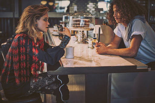 Two people sit at a marble counter in a diner. An adult female holds glasses and wears a fringed top. An adult male is visible across from her.