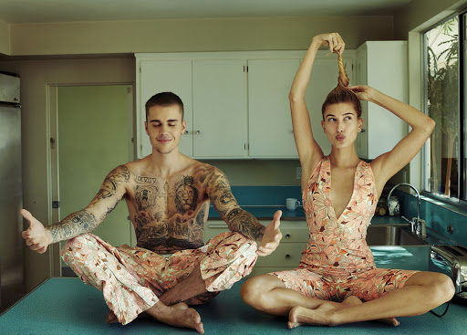 Justin and Hailey Bieber pose in matching floral pajamas in a kitchen, sitting cross-legged on a blue countertop with white cabinets.
