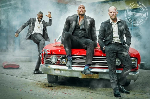 Three men in suits lean on a red car amidst a smoky scene. Dwayne Johnson, Jason Statham, and Idris Elba pose for an Entertainment Weekly cover.