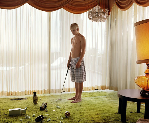 A young, shirtless man in plaid shorts stands on a green carpet, appearing to have just played indoor golf with broken vases nearby.
