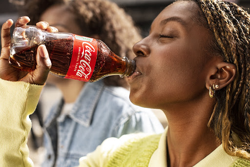 Young African American woman drinks Coca-Cola from a glass bottle outdoors in daylight, wearing a lime green sweater, medium shot.