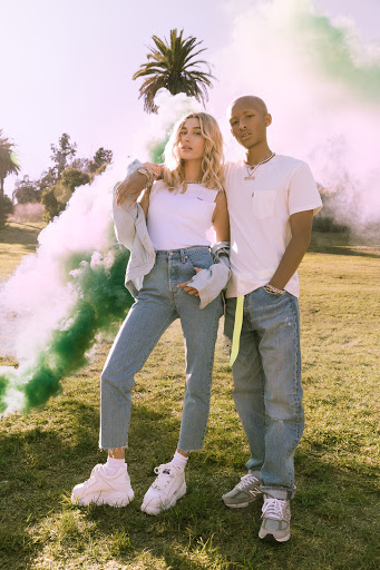 Two young adults pose in a grassy field with green smoke, wearing casual clothing and sneakers, under bright sunlight with trees nearby.