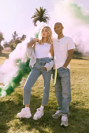 Two young adults pose in a grassy field with green smoke, wearing casual clothing and sneakers, under bright sunlight with trees nearby.