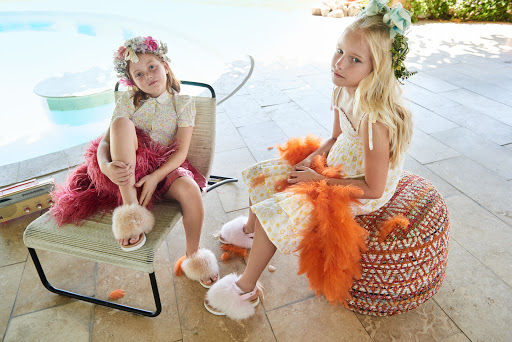Two young girls in floral dresses and flower crowns sit outdoors near a pool, one on a chair and the other on an ottoman.