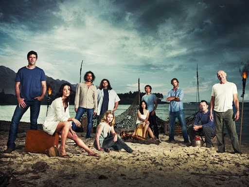 Group of people on a sandy beach with water, mountains, and cloudy sky in the background. Some are sitting around a fire.