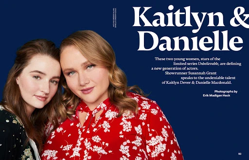 Close-up of Kaitlyn Dever and Danielle Macdonald posing in a photo studio with blue backdrop. Dever wears floral-print dress, Macdonald wears dark dress.
