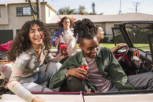 Group of four young adults smile while sitting in a convertible, each holding a Coca-Cola bottle on a sunny day in a suburban neighborhood.