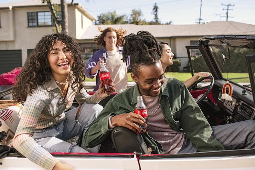 Group of four young adults smile while sitting in a convertible, each holding a Coca-Cola bottle on a sunny day in a suburban neighborhood.