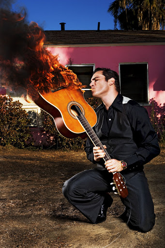 A man in a black suit crouches while holding a flaming guitar with smoke, set against a pink house and clear blue sky backdrop.