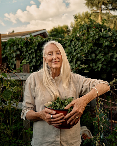 Elderly woman poses outdoors in a garden with a bowl of leafy greens, wearing a light linen shirt and rings. Natural light.